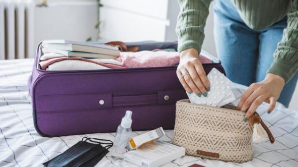 Woman preparing travel suitcase on bed at home. She is packing medicine and disinfection stuff for travel.
